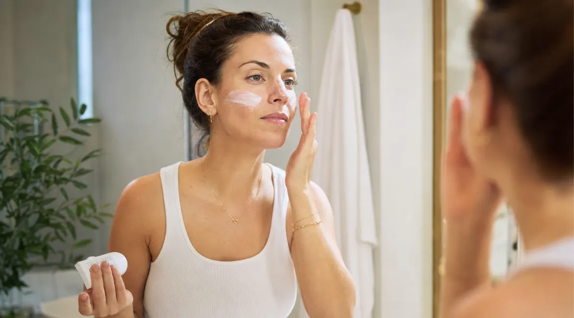 A woman applying sunscreen to her face in natural morning light near a bright window