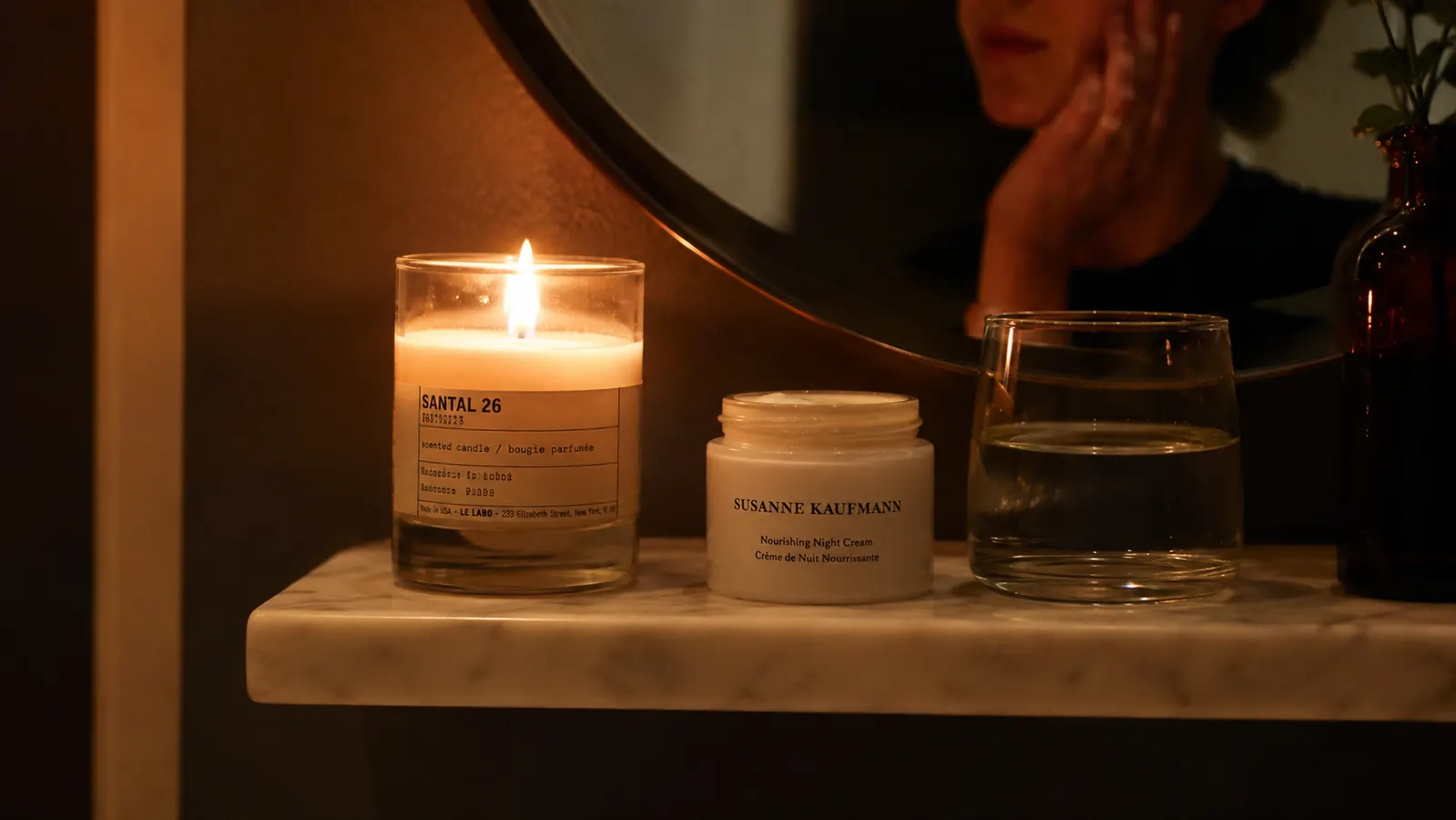 Close-up of a bathroom shelf at night with a small white jar of RoC Retinol Correxion beside a glass of water and a softly lit candle